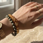 Hand wearing multiple bracelets pf pyrite and citrine and rings on a neutral background