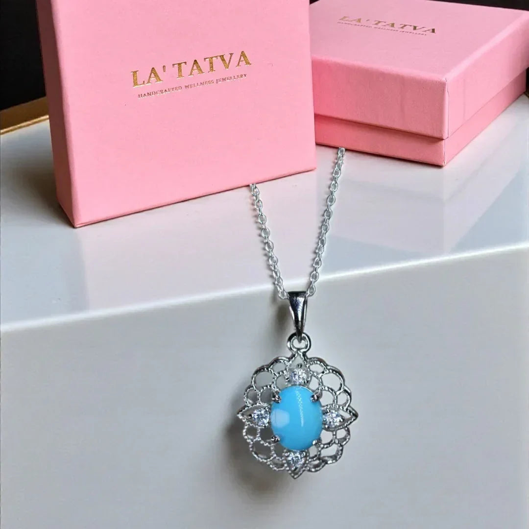 Turquoise pendant necklace with silver chain on a white surface, against a pink 'LA' TATVA' box.
